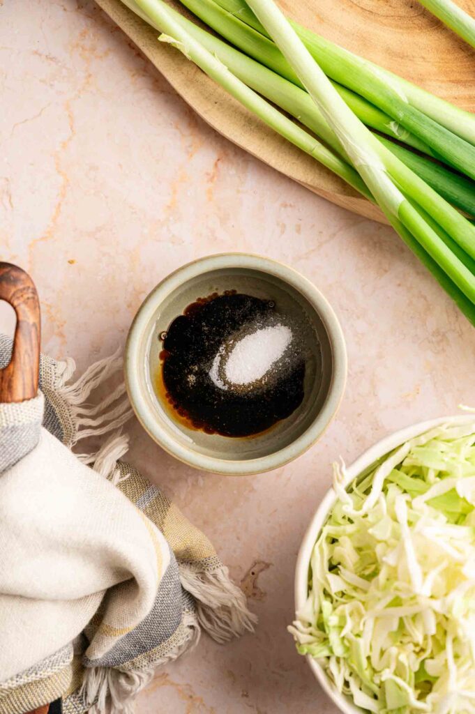 A small bowl of sauce with seasonings sits on a countertop beside shredded cabbage, whole green onions, and a striped cloth—classic ingredients ready to create Panda Express Chow Mein at home.