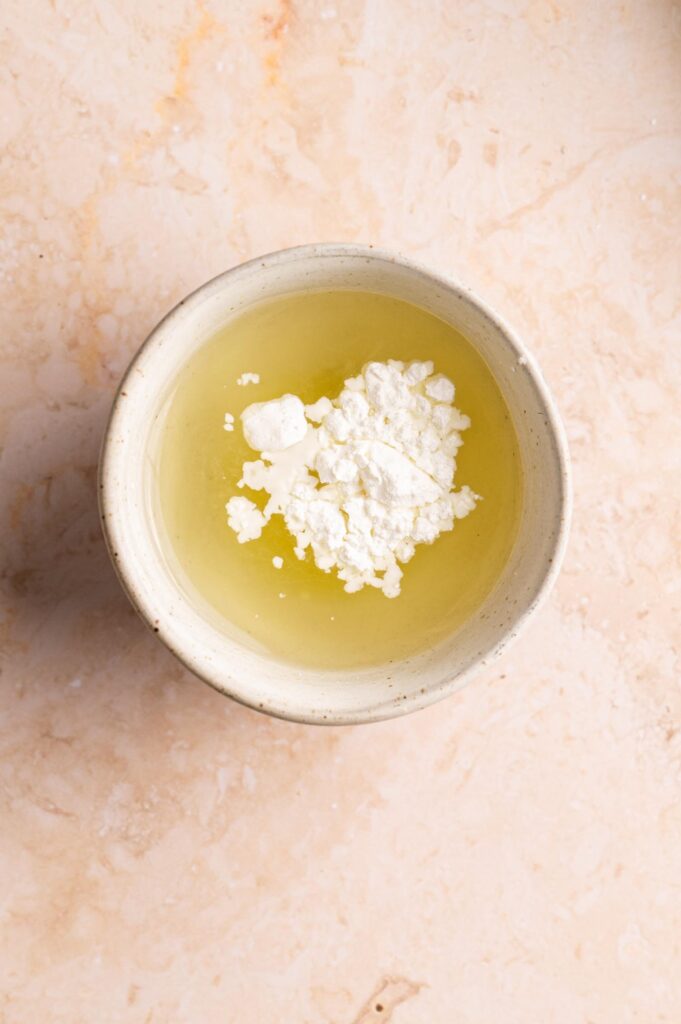 A ceramic bowl containing egg whites and a small pile of white powder, likely cream of tartar, sits on a light-colored marble surface—an essential step before crafting dishes that call for perfectly whipped egg whites, much like those found in some homemade versions of Panda Express Chow Mein.