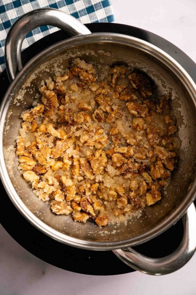 Chopped walnuts coated in a crystallized sugar mixture, just like the topping on Panda Express Honey Walnut Shrimp, in a stainless steel pot on a stove, with a checkered cloth partially visible.