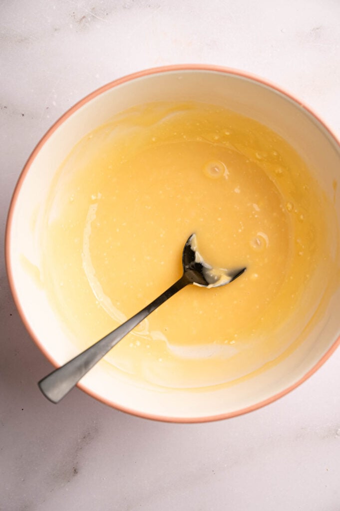 A metal spoon rests in a bowl containing a smooth, pale yellow mixture reminiscent of Panda Express Honey Walnut Shrimp sauce, set on a light-colored surface.