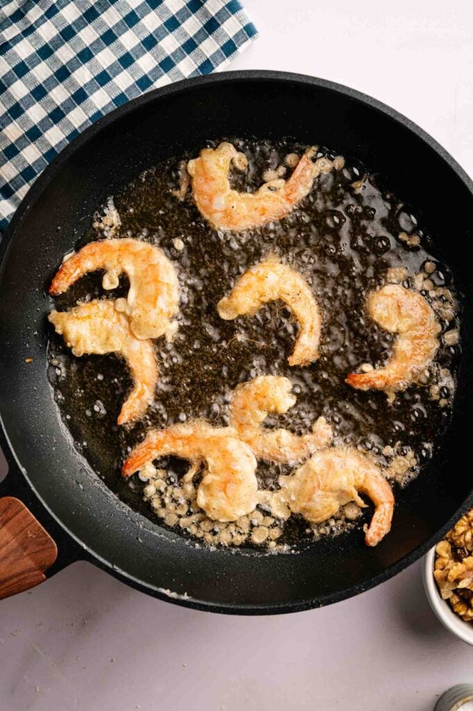 Shrimp coated in batter, reminiscent of Panda Express Honey Walnut Shrimp, are being deep-fried in oil in a black skillet; a blue and white checkered cloth is nearby.