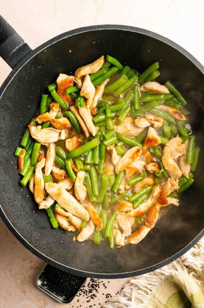 A stir fry in a black pan with cooked chicken strips and green beans in a light sauce, reminiscent of Panda Express String Bean Chicken Breast.