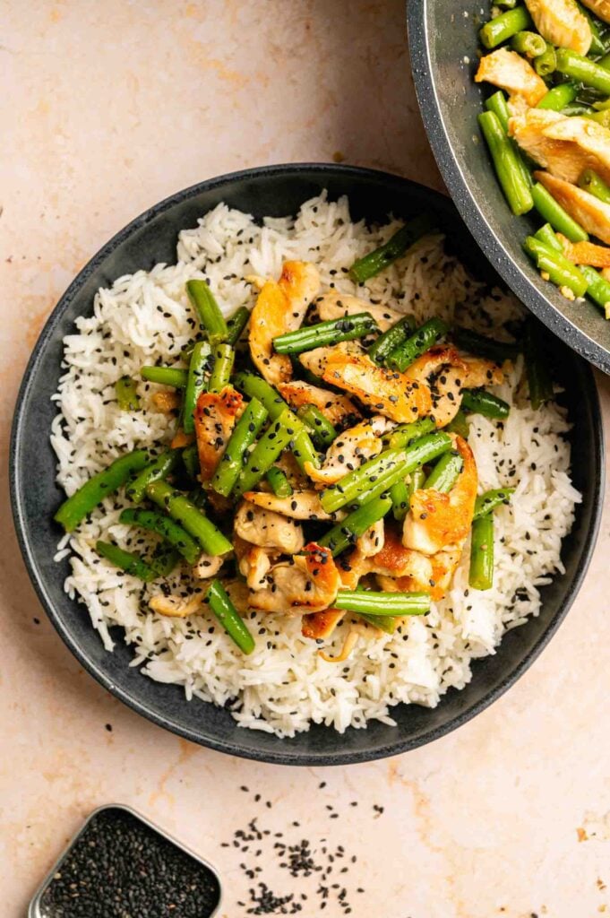 A bowl of white rice topped with stir-fried chicken, green beans, and black sesame seeds, reminiscent of Panda Express String Bean Chicken Breast, with a pan of the same stir-fry partially visible above.
