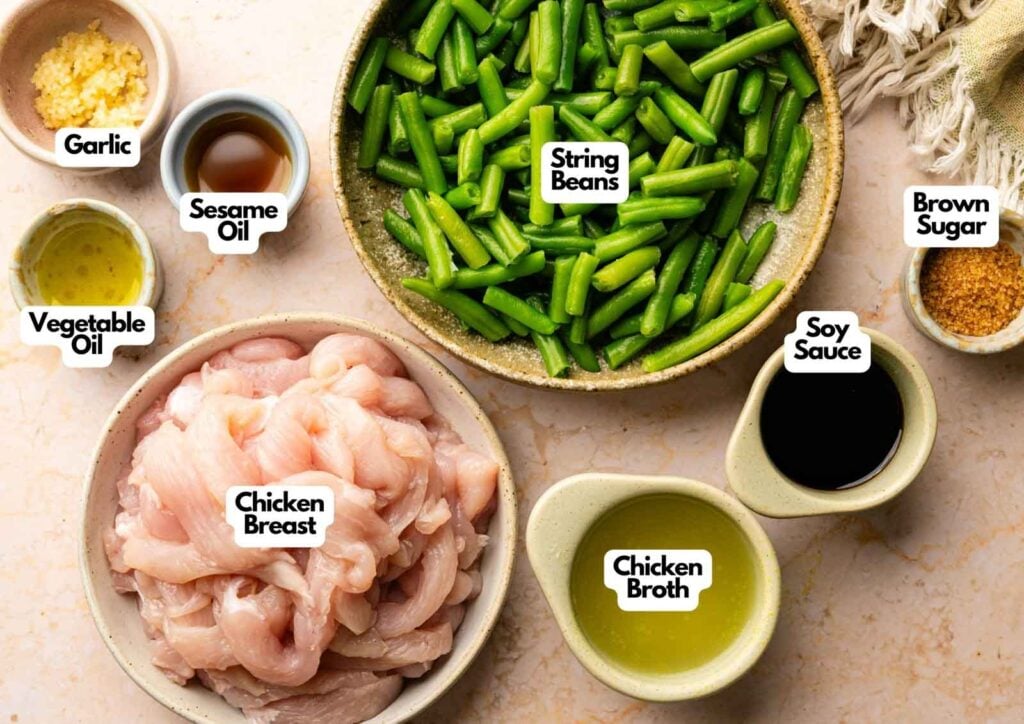 Bowls containing ingredients for Panda Express String Bean Chicken Breast&mdash;sliced chicken breast, chopped string beans, soy sauce, chicken broth, sesame oil, vegetable oil, garlic, and brown sugar&mdash;are all labeled on a countertop.