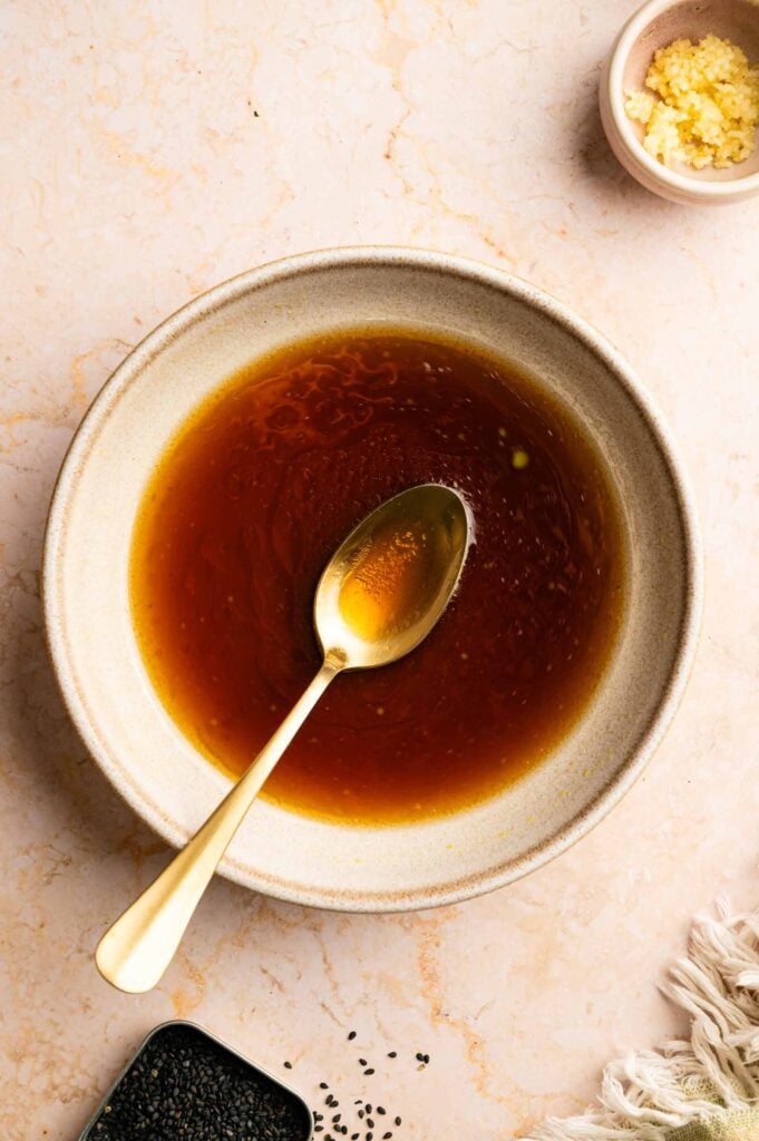 A bowl of brown liquid sauce with a spoon resting inside, surrounded by a small bowl of minced garlic and scattered black sesame seeds, reminiscent of the flavors found in Panda Express String Bean Chicken Breast, on a light-colored surface.
