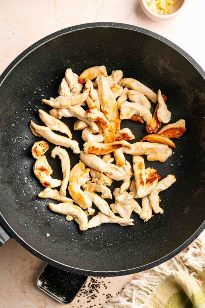 Strips of chicken cooking and browning in a black wok, reminiscent of Panda Express String Bean Chicken Breast, with chopped garlic and a small bowl of sesame seeds visible nearby.