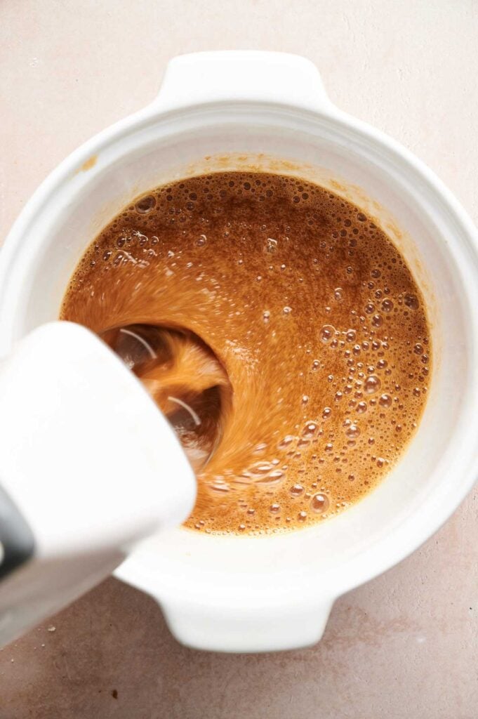 A white electric mixer blends a frothy brown liquid in a white mixing bowl on a light-colored surface, preparing the filling for delicious Air Fryer Pecan Pie Bars.