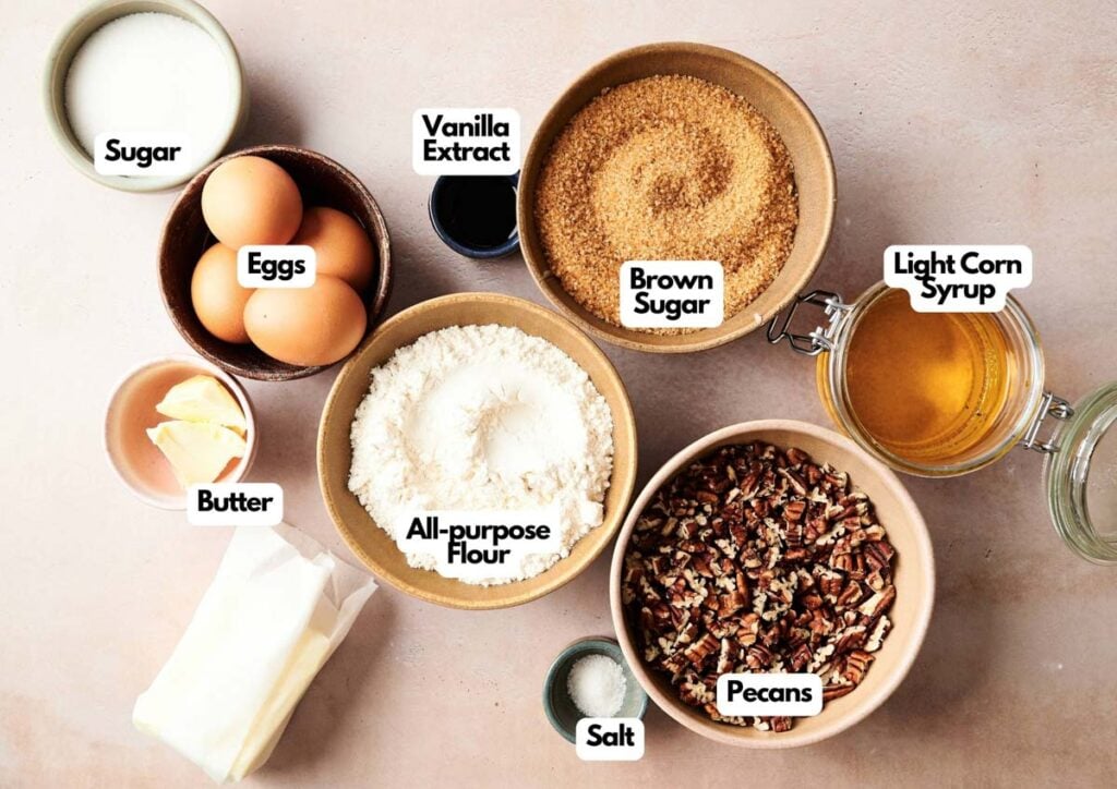 A top-down view of labeled baking ingredients in bowls and jars for Air Fryer Pecan Pie Bars: sugar, eggs, butter, all-purpose flour, vanilla extract, brown sugar, light corn syrup, pecans, and salt.