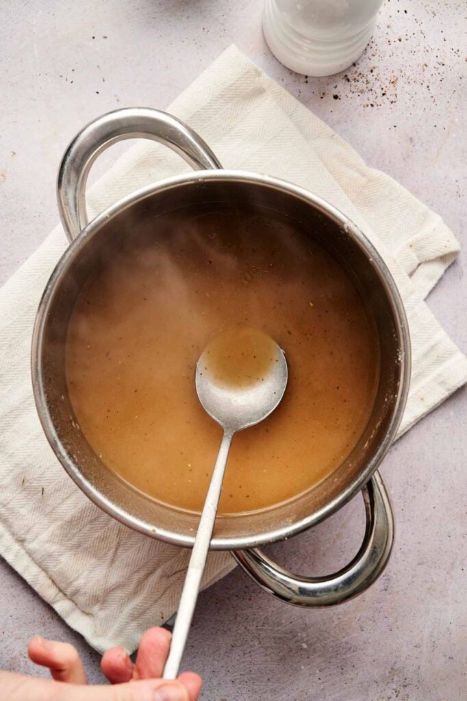 A hand holds a ladle in a pot of brown gravy on a white towel, with steam rising from the surface—perfect for serving over Air Fryer Turkey Meatballs.