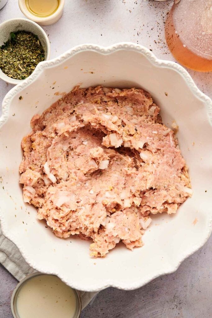 A white bowl filled with raw ground meat mixture, perfect for making Air Fryer Turkey Meatballs, is surrounded by small bowls of herbs and seasonings on a light countertop.