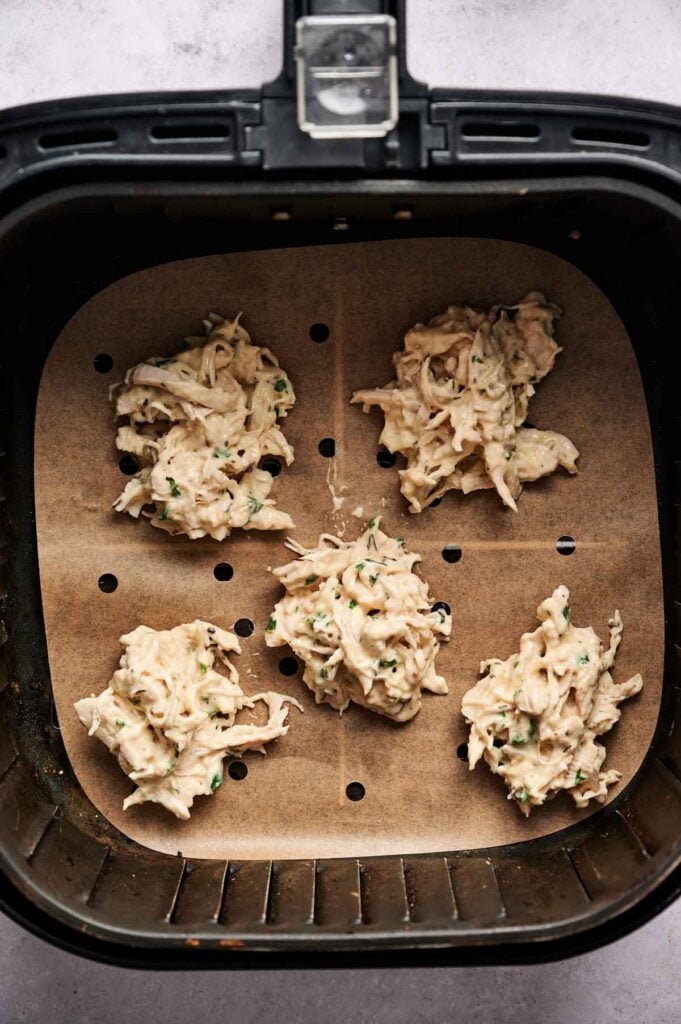 Five raw Air Fryer Chicken Fritters, made with shredded ingredients and herbs, are placed on parchment paper inside an air fryer basket, ready to be cooked.