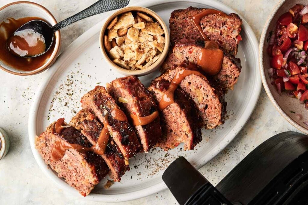 Sliced Air Fryer Cracker Barrel Meatloaf with sauce drizzled on top, served on a white plate with a bowl of croutons and a side dish of chopped tomatoes and onions.