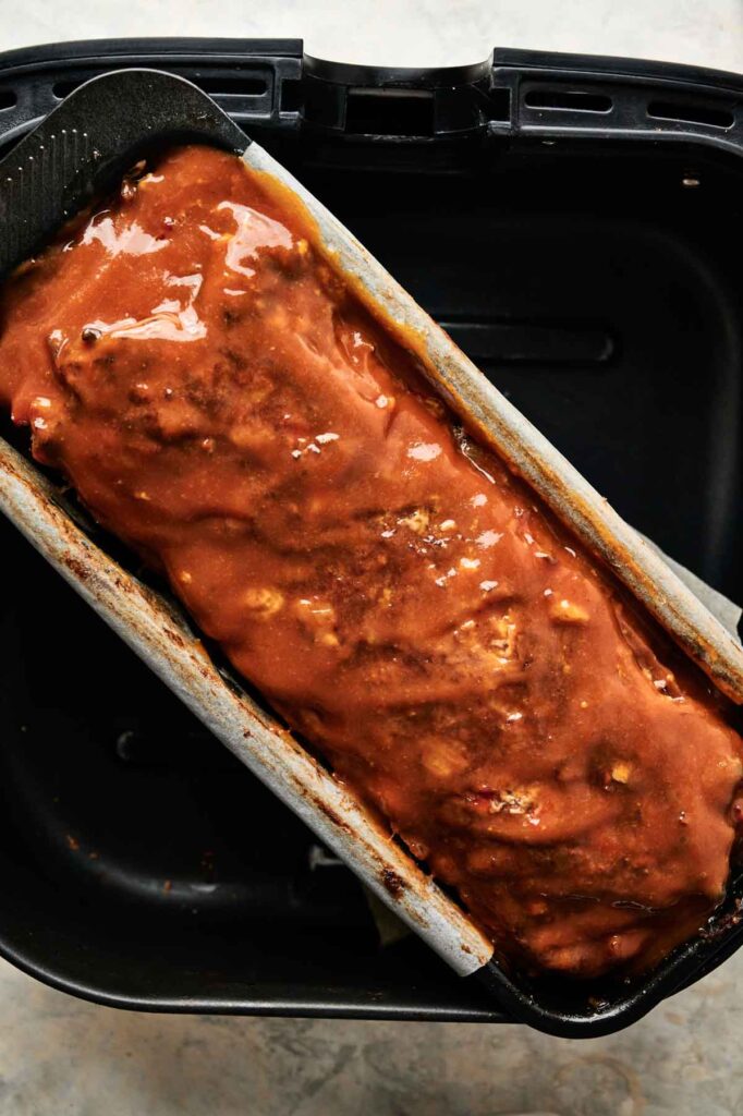 An Air Fryer Cracker Barrel Meatloaf covered in sauce sits in a rectangular pan inside an air fryer basket.