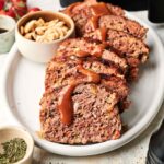 Sliced Air Fryer Cracker Barrel Meatloaf with brown sauce on a white oval plate, served alongside a small bowl of cashews and a dish of dried herbs.