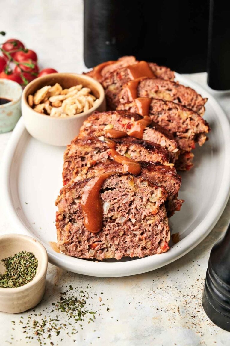 Sliced Air Fryer Cracker Barrel Meatloaf with brown sauce on a white oval plate, served alongside a small bowl of cashews and a dish of dried herbs.