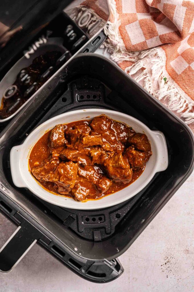 A white oval dish filled with saucy Air Fryer Lamb Rogan Josh sits inside an air fryer basket, with a pink and white towel nearby on the counter.