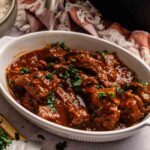 Oval white dish with Air Fryer Lamb Rogan Josh pieces in a thick, red sauce, garnished with chopped herbs; a bowl of rice and a knife with herbs are nearby.