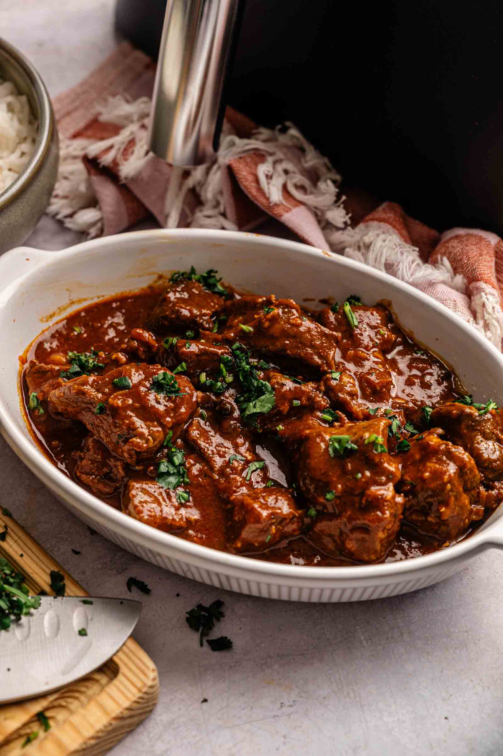 Oval white dish with Air Fryer Lamb Rogan Josh pieces in a thick, red sauce, garnished with chopped herbs; a bowl of rice and a knife with herbs are nearby.