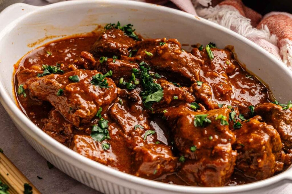 Beef pieces in a rich, reddish-brown sauce reminiscent of Air Fryer Lamb Rogan Josh, garnished with chopped herbs and served in a white oval dish.