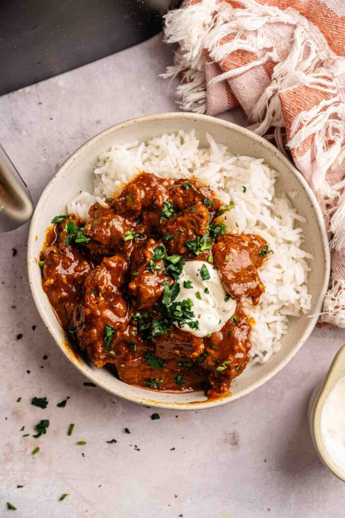 A bowl of white rice topped with Air Fryer Lamb Rogan Josh, garnished with chopped herbs and a dollop of sour cream, placed on a light surface next to a pink cloth napkin.