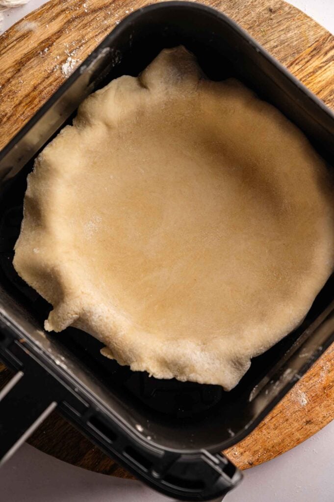 Unbaked pie crust placed in a square black baking pan, set on a round wooden board—perfect for preparing an Air Fryer Marry Me Chicken Pot Pie.