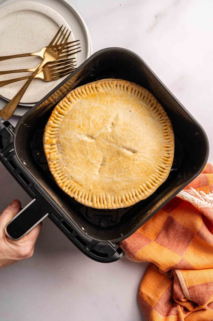 A hand holds an air fryer basket containing a baked Air Fryer Marry Me Chicken Pot Pie. Plates, forks, and an orange checked cloth are arranged nearby on a white surface.