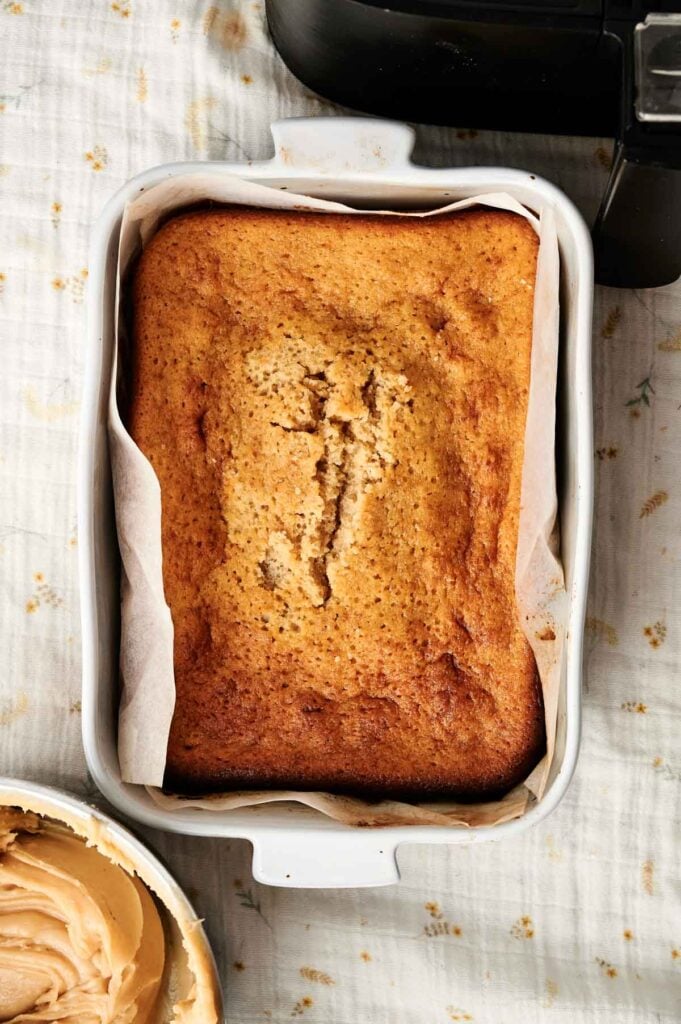 A rectangular loaf of air-fryer peanut butter cake in a parchment-lined white dish, set on a light-patterned surface next to a bowl of peanut butter.