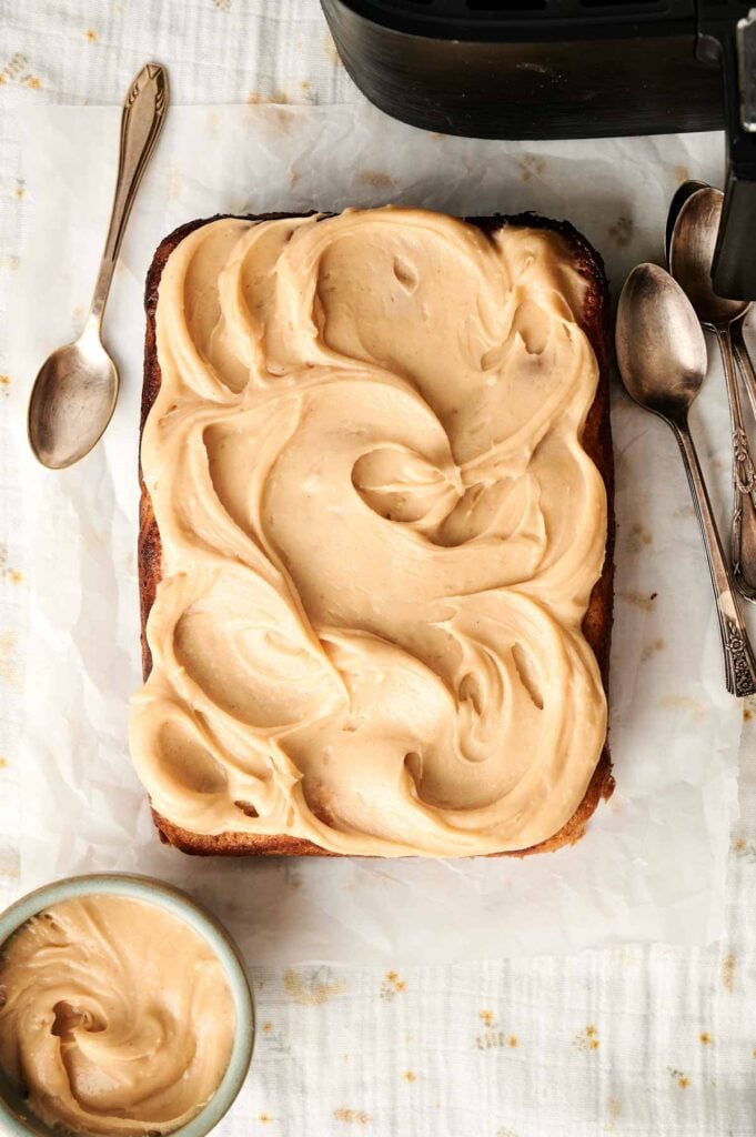 An Air Fryer Peanut Butter Cake, rectangular and topped with swirled light brown frosting, sits on parchment paper beside several metal spoons and a small bowl of extra frosting.