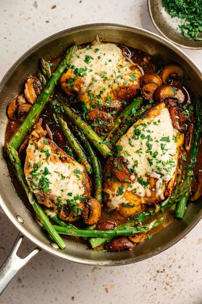 Chicken breasts topped with melted cheese, surrounded by asparagus and mushrooms in a skillet, garnished with chopped parsley&mdash;a classic inspired by Cheesecake Factory Chicken Madeira.