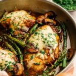 Three cooked chicken breasts topped with melted cheese, mushrooms, and chopped herbs in a skillet with asparagus and savory sauce—a homestyle take reminiscent of Cheesecake Factory Chicken Madeira—with a bowl of chopped parsley in the background.