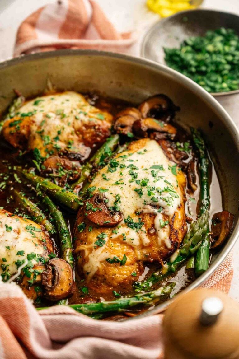 Three cooked chicken breasts topped with melted cheese, mushrooms, and chopped herbs in a skillet with asparagus and savory sauce—a homestyle take reminiscent of Cheesecake Factory Chicken Madeira—with a bowl of chopped parsley in the background.
