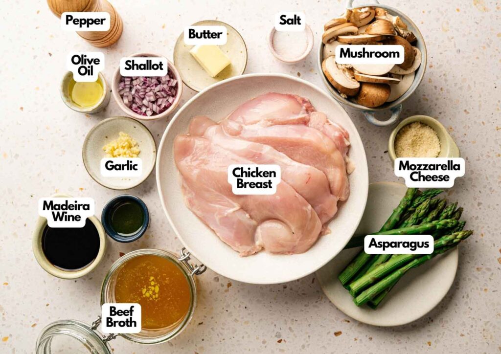 Top-down view of labeled ingredients in bowls and plates for Cheesecake Factory Chicken Madeira, featuring chicken breast, asparagus, mushrooms, mozzarella cheese, butter, shallot, olive oil, garlic, Madeira wine, beef broth, pepper, and salt.