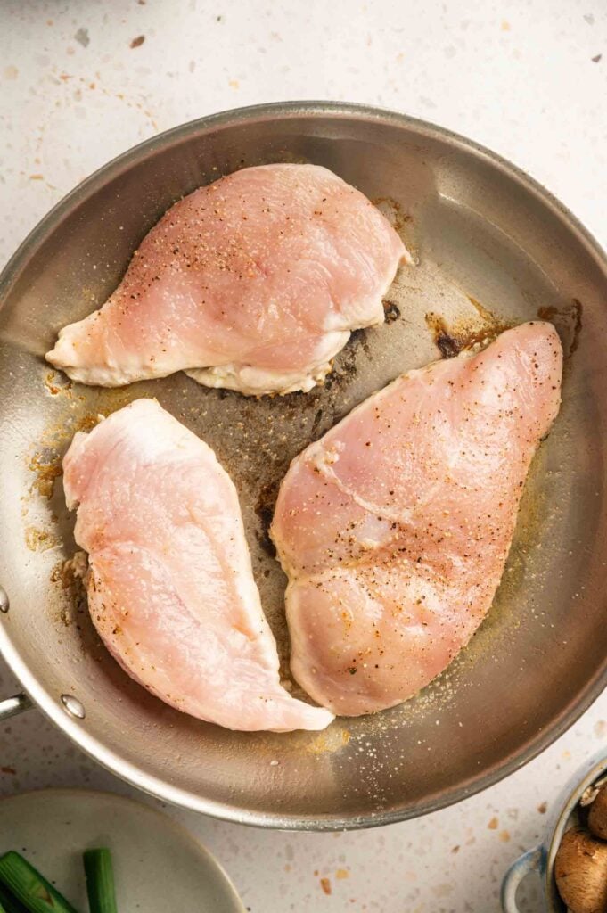 Seasoned chicken breasts searing in a stainless steel skillet.