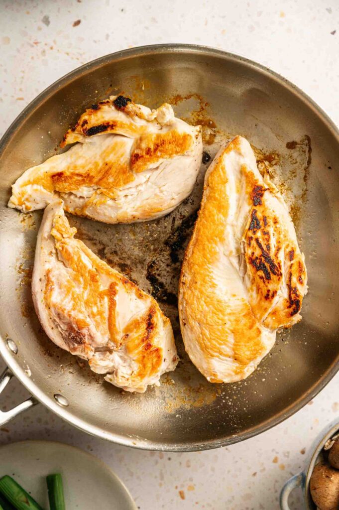 Three pieces of seared chicken breast, inspired by Cheesecake Factory Chicken Madeira, cook in a stainless steel skillet on a light-colored countertop.