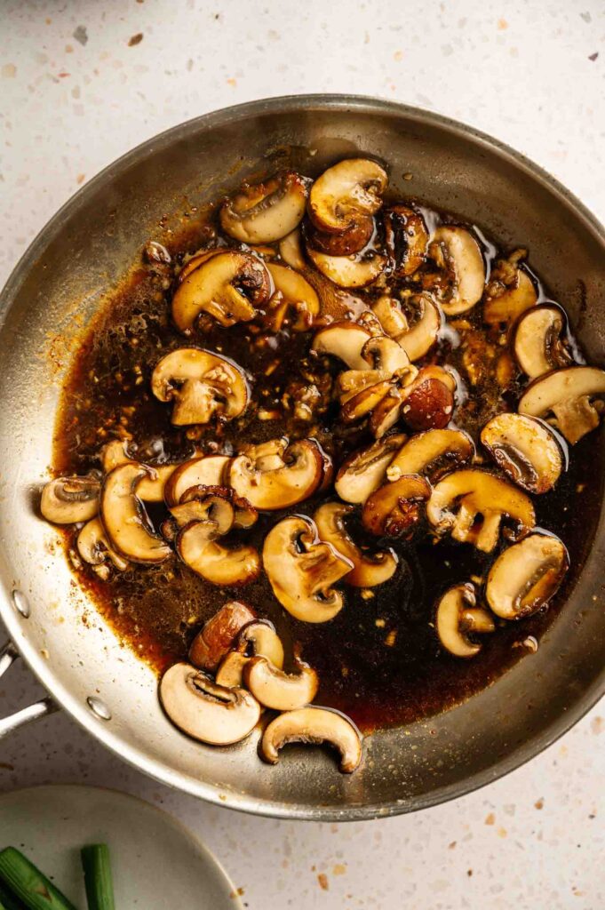 Sliced mushrooms cooking in a stainless steel pan with a dark, savory sauce, just like the ones in Cheesecake Factory Chicken Madeira, on a light-colored countertop.