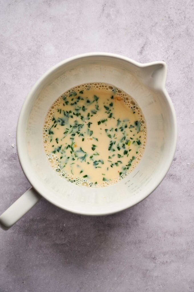 A white mixing bowl containing a beaten egg mixture with chopped herbs, perfect for preparing Air Fryer Chicken Fritters, sits on a light gray surface.