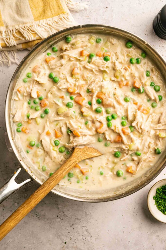 A skillet filled with creamy Chicken Pot Pie with Cheddar Bay Biscuits, featuring a savory chicken and vegetable mixture—peas and carrots included—with a wooden spoon resting inside.
