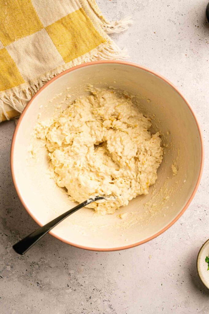 A mixing bowl with a spoon contains thick, lumpy Chicken Pot Pie with Cheddar Bay Biscuits batter on a light-colored countertop next to a yellow and white checkered cloth.