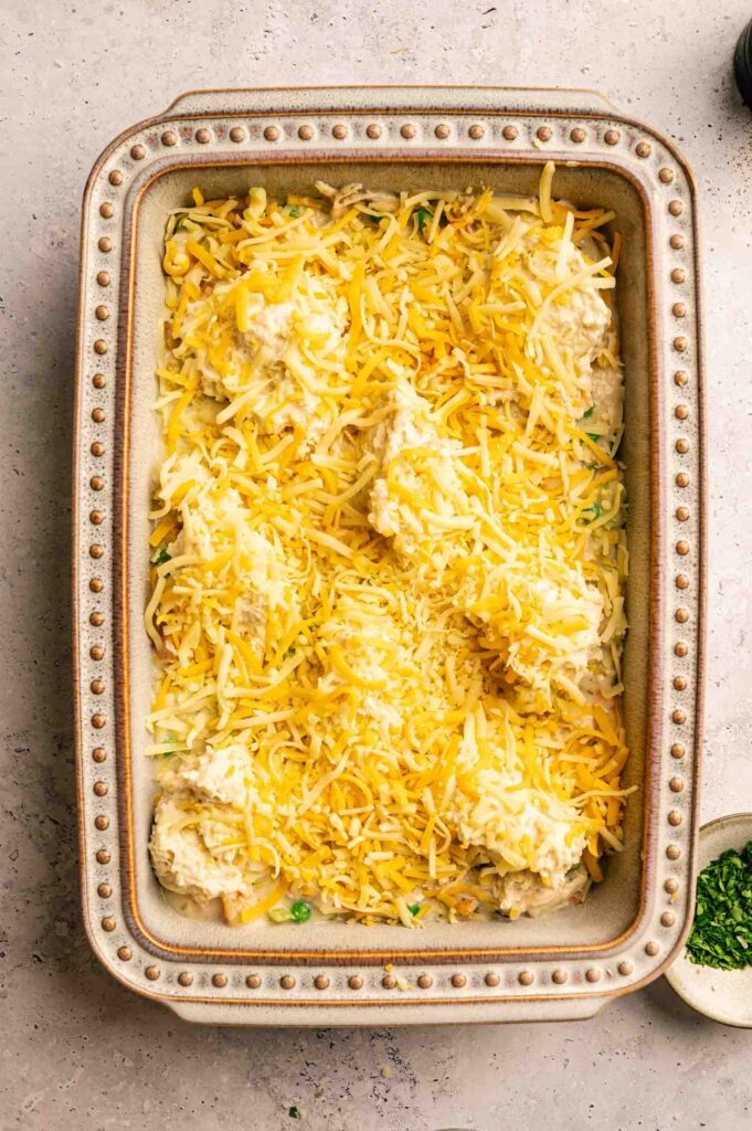 A rectangular baking dish filled with an unbaked Chicken Pot Pie with Cheddar Bay Biscuits, generously topped with shredded cheese, sits on a light-colored surface.