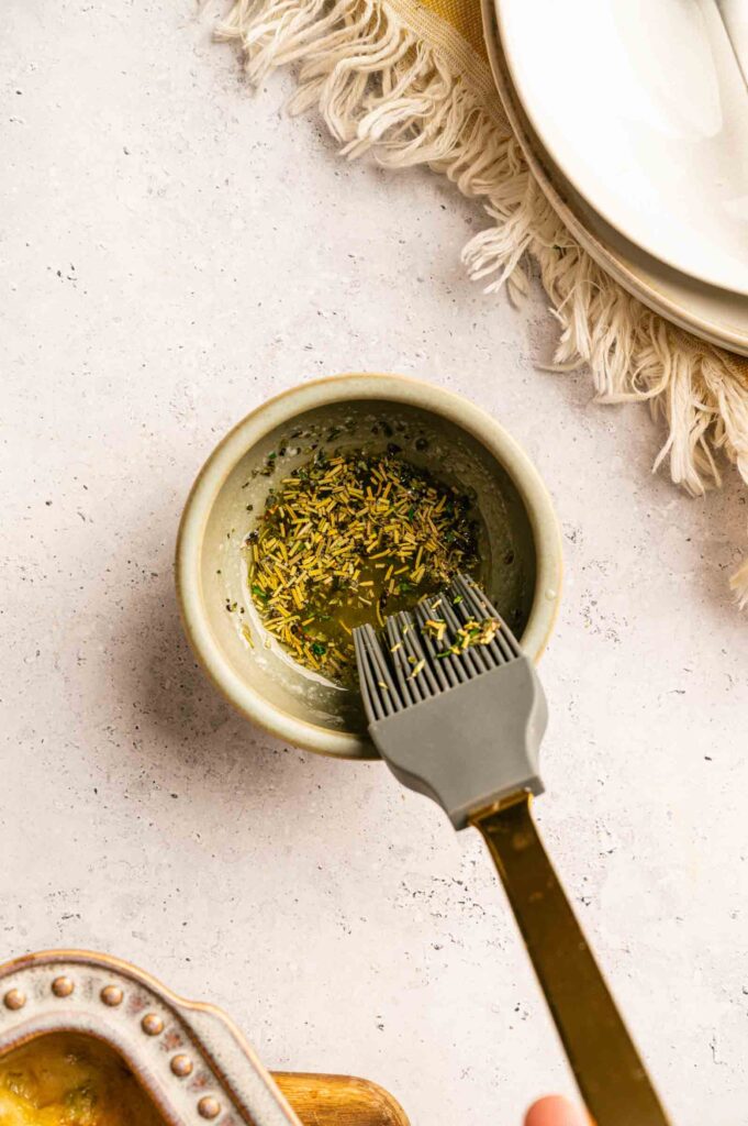 A bowl of seasoned butter with a basting brush rests inside, set on a light countertop near stacked plates and a fringed napkin—an inviting setup for preparing Chicken Pot Pie with Cheddar Bay Biscuits.