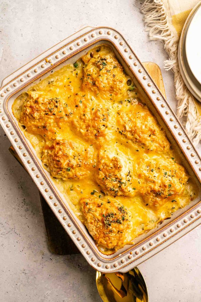 A rectangular baking dish filled with Chicken Pot Pie with Cheddar Bay Biscuits, topped with melted cheese and herbs, sits on a countertop with a spoon and a fringed napkin nearby.