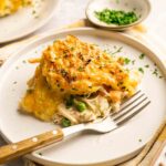 A serving of Chicken Pot Pie with Cheddar Bay Biscuits features a golden, cheesy crust and a savory filling of peas and chicken, garnished with chopped herbs on a white plate with a fork.