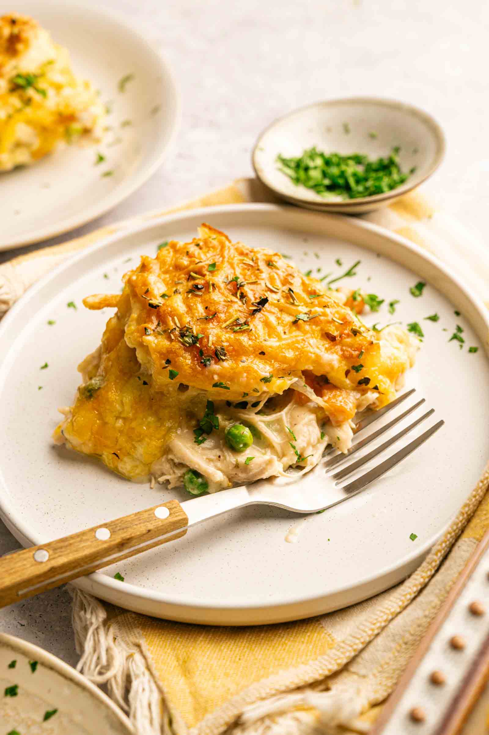 A serving of Chicken Pot Pie with Cheddar Bay Biscuits features a golden, cheesy crust and a savory filling of peas and chicken, garnished with chopped herbs on a white plate with a fork.