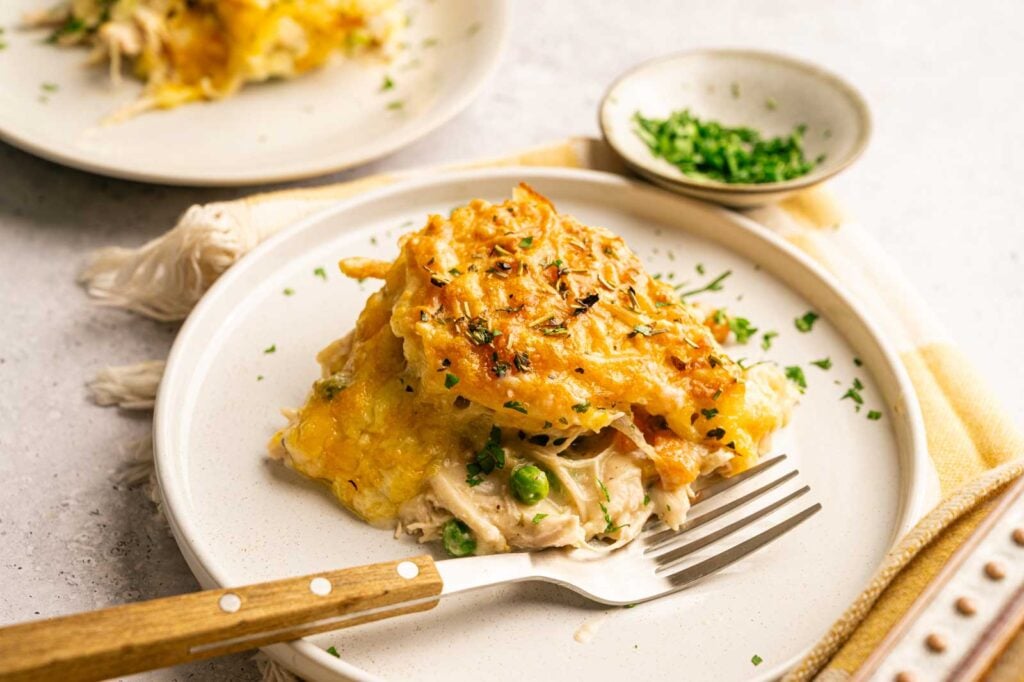 A serving of Chicken Pot Pie with Cheddar Bay Biscuits, featuring a golden baked cheese topping and peas on a white plate, garnished with chopped herbs, with a fork on the side.