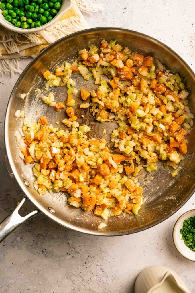 Chopped onions, celery, and carrots sauté in a large metal skillet on a light countertop—essential flavors for Chicken Pot Pie with Cheddar Bay Biscuits, with bowls of peas, chives, and cream ready to add richness.