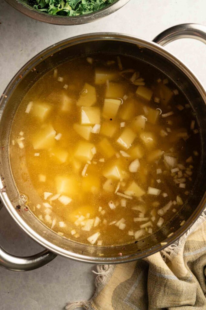 A pot filled with chopped potatoes and onions simmering in broth, inspired by Copycat Olive Garden Zuppa Toscana, with a kitchen towel and a bowl of greens nearby.