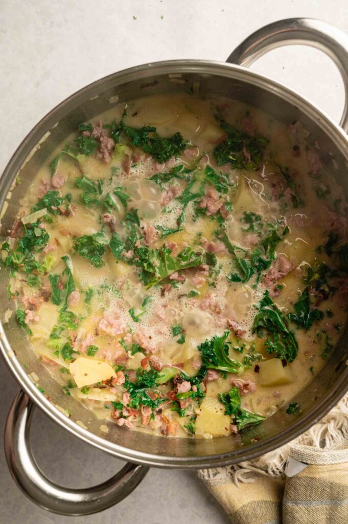 A large pot of Copycat Olive Garden Zuppa Toscana soup with kale, potatoes, sausage, and broth sits next to a beige and gray striped cloth on a light surface.