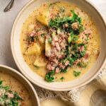 A bowl of creamy Copycat Olive Garden Zuppa Toscana, filled with potatoes, sausage, and kale, is garnished with grated cheese and red pepper flakes, sitting on a table next to a spoon and a small bowl of grated cheese.