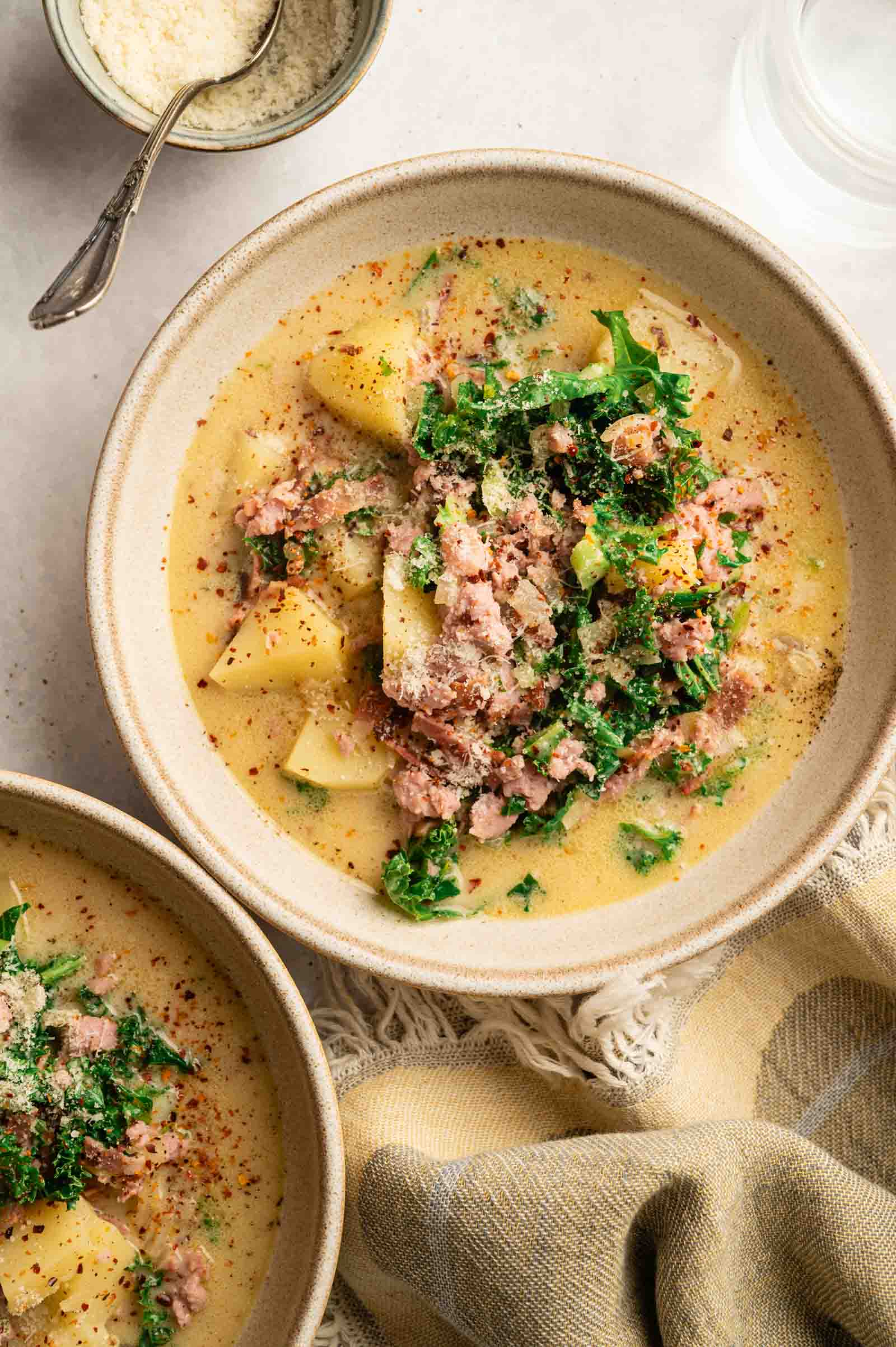 A bowl of creamy Copycat Olive Garden Zuppa Toscana, filled with potatoes, sausage, and kale, is garnished with grated cheese and red pepper flakes, sitting on a table next to a spoon and a small bowl of grated cheese.