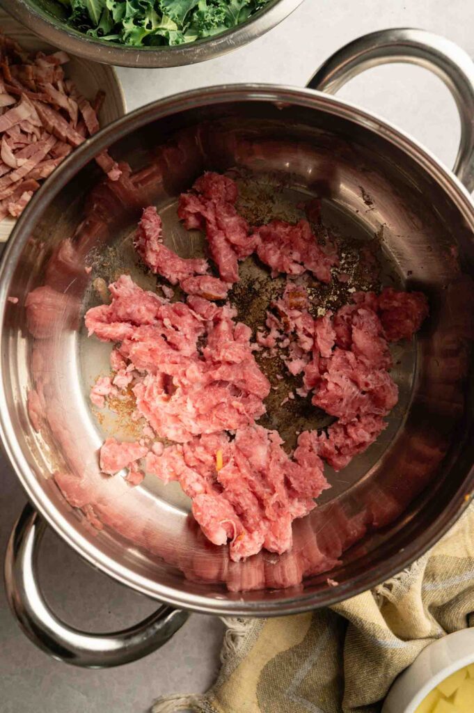 Raw Italian sausage is scattered in a stainless steel pot, with bowls of chopped greens and sliced meat nearby on a light countertop—perfect prep for a Copycat Olive Garden Zuppa Toscana.